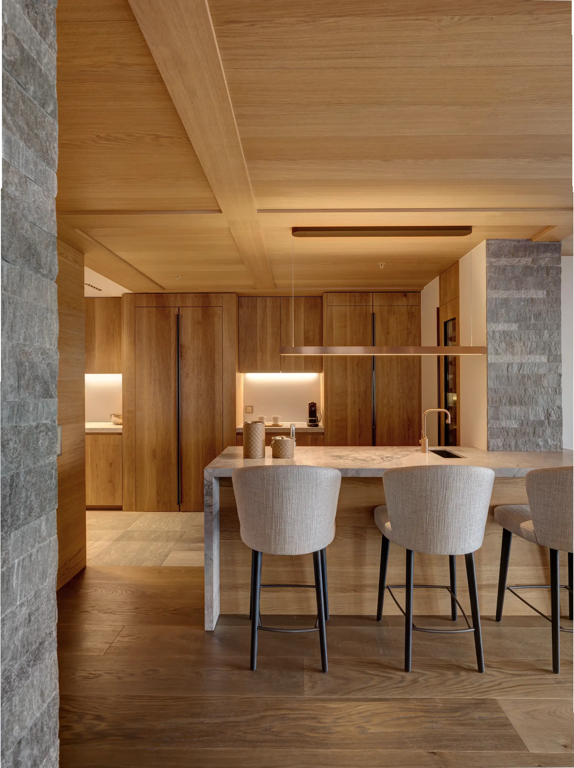 Kitchen island and dining counter with upholstered bar stools, set between brushed timber cabinetry and rough granite-clad columns under a coffered wood ceiling
