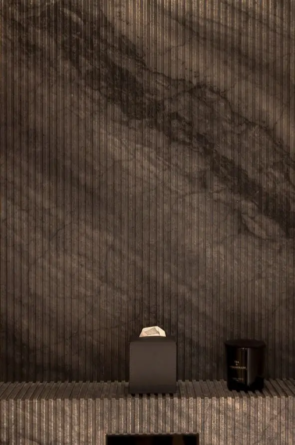 Detail of a dark veined stone wall above a fluted stone shelf displaying a tissue box and a black scented candle