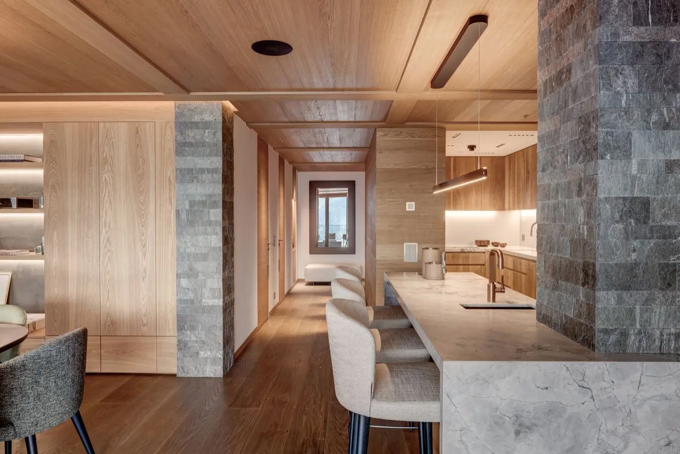 Wide view of the living and kitchen space showing a marble island, granite-clad columns, timber ceiling and a corridor framing a mountain view through a recessed window